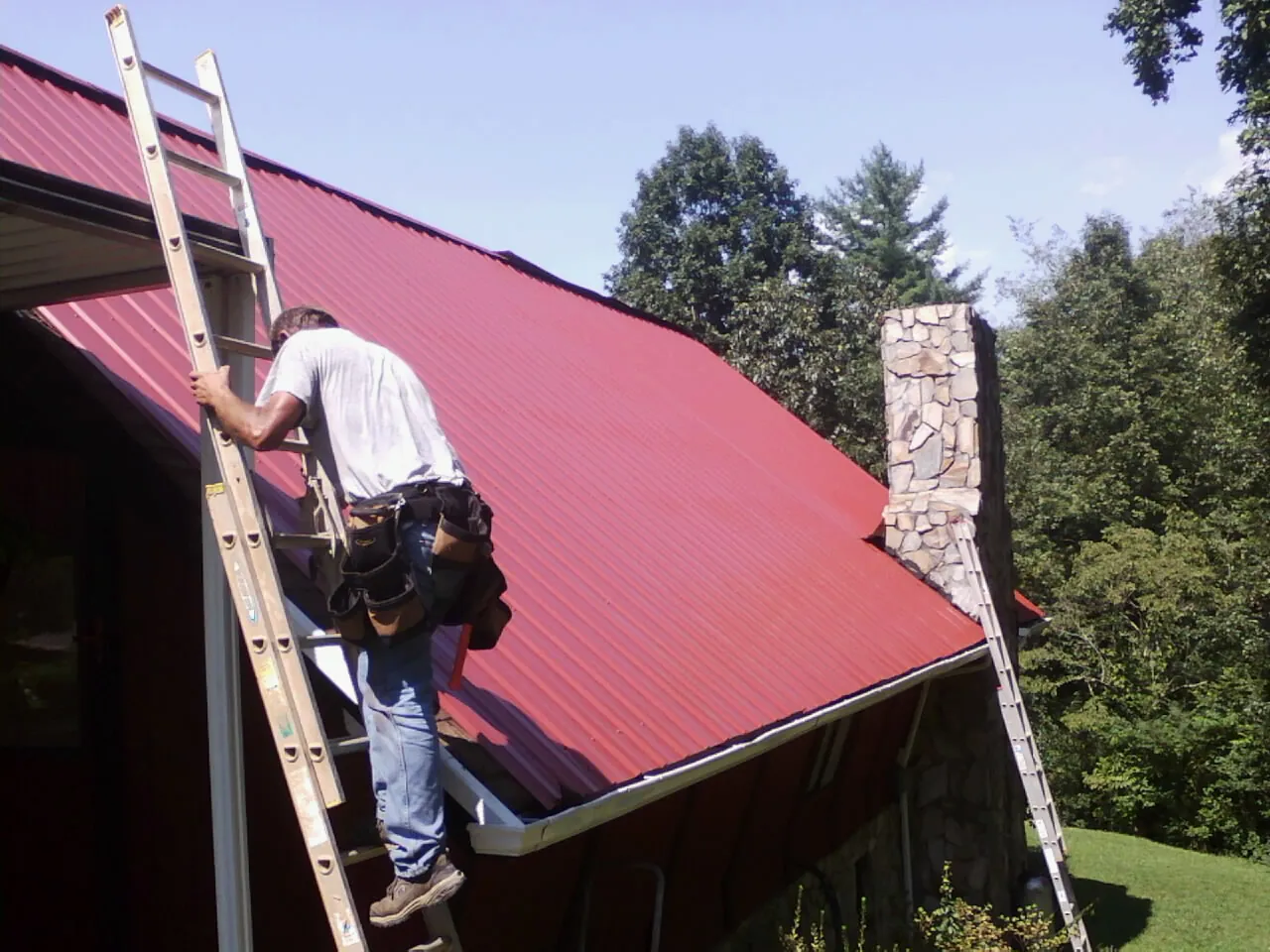 Roofer on a ladder beside a red metal roof and stone chimney — Soffit & Fascia Repair by Roofing Brothers in Dillard